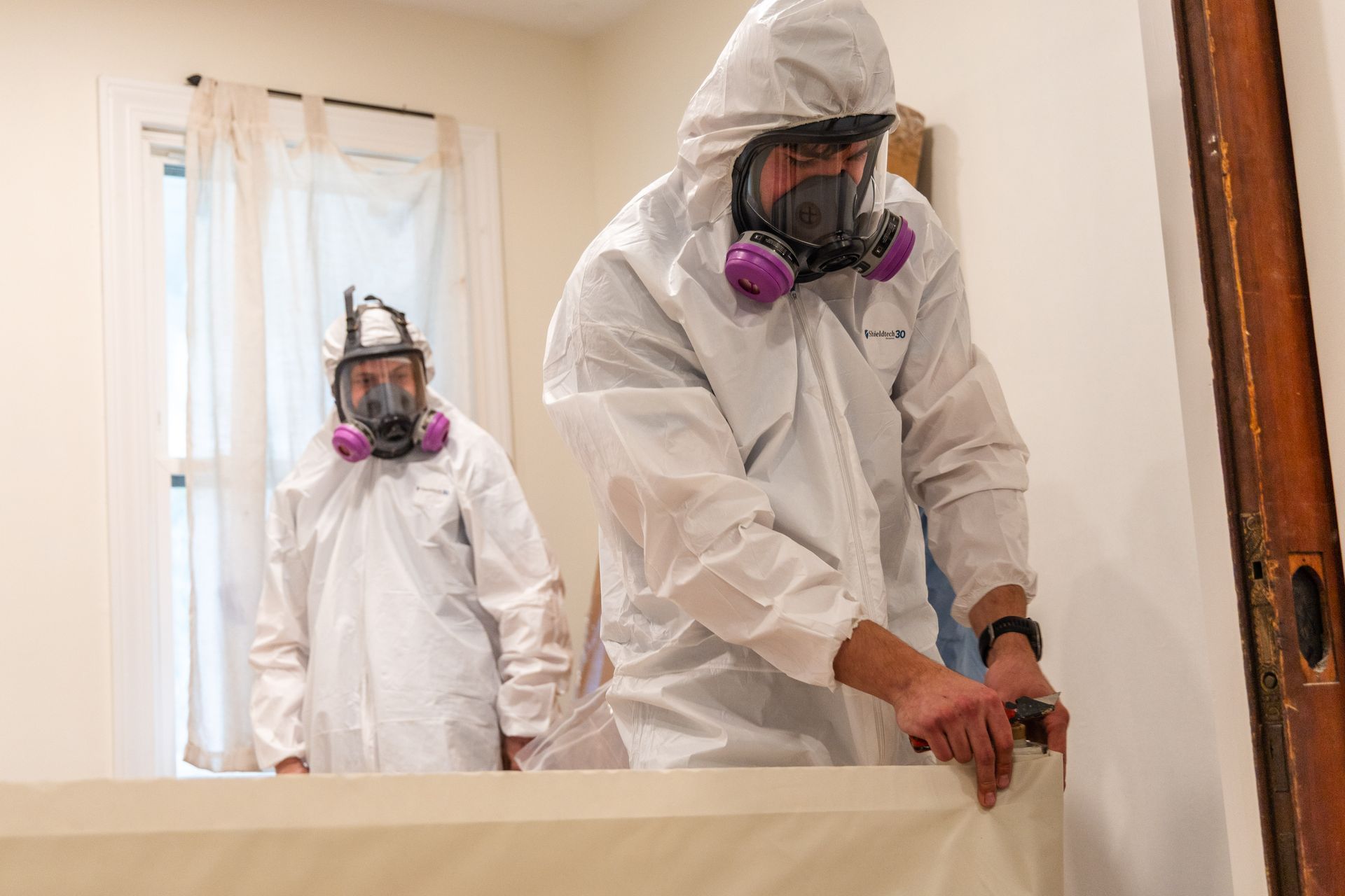 Two workers in hazmat suits and respirators cleaning a room with a window and mirror.