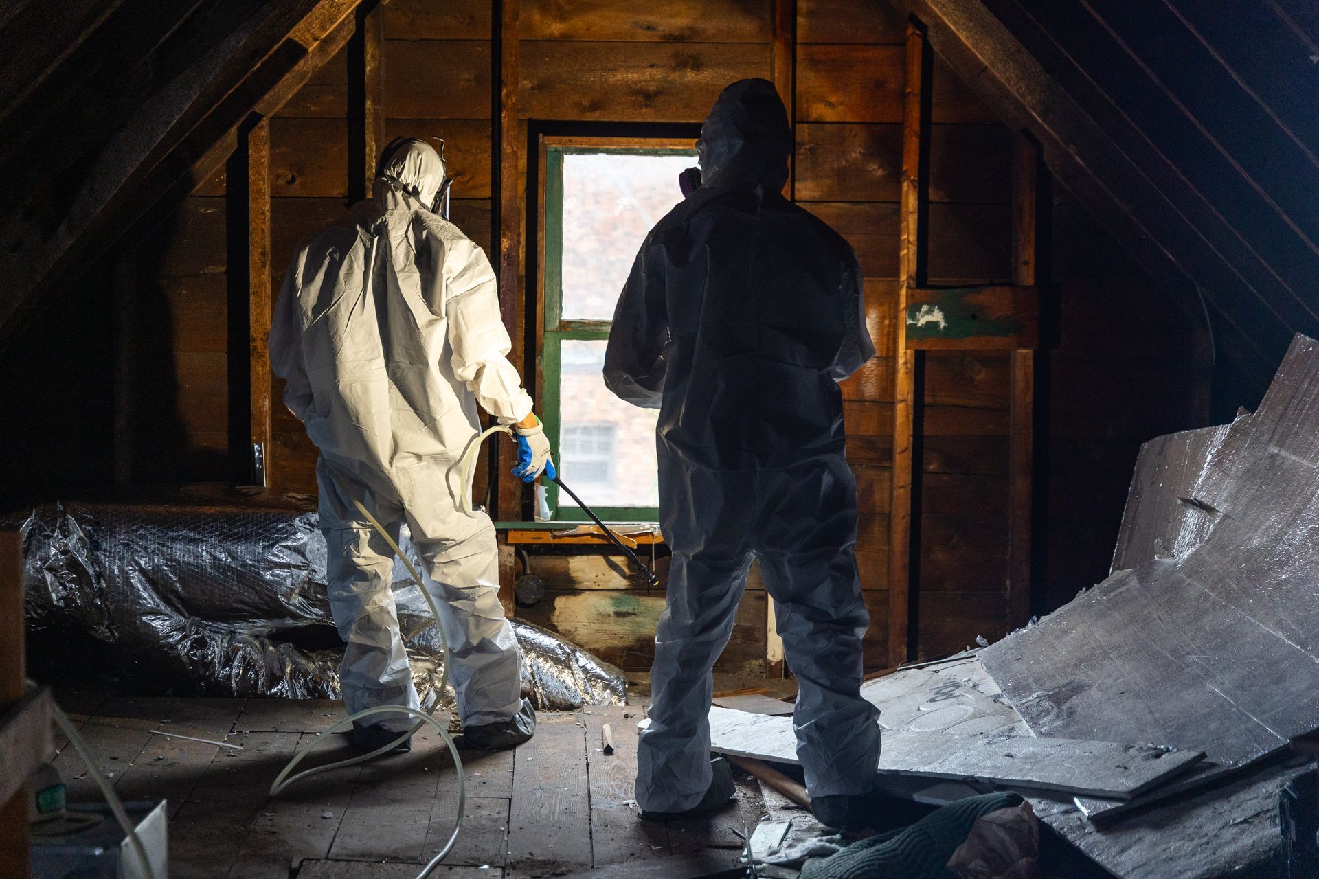Two people in protective suits inspect a damaged attic with insulation debris and a bright window.