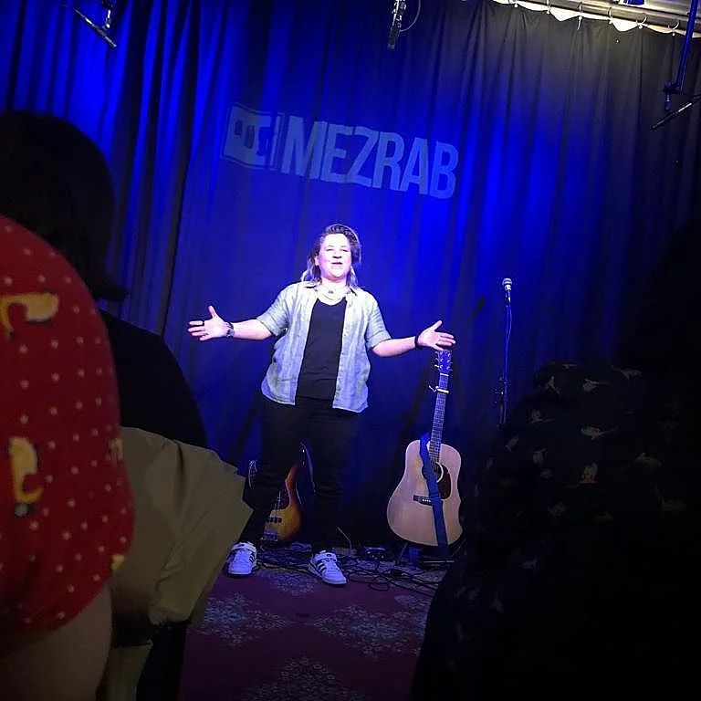 Artiest op een klein podium met uitgestrekte armen onder blauwe lichten, gitaar in de buurt, achtergrond met
