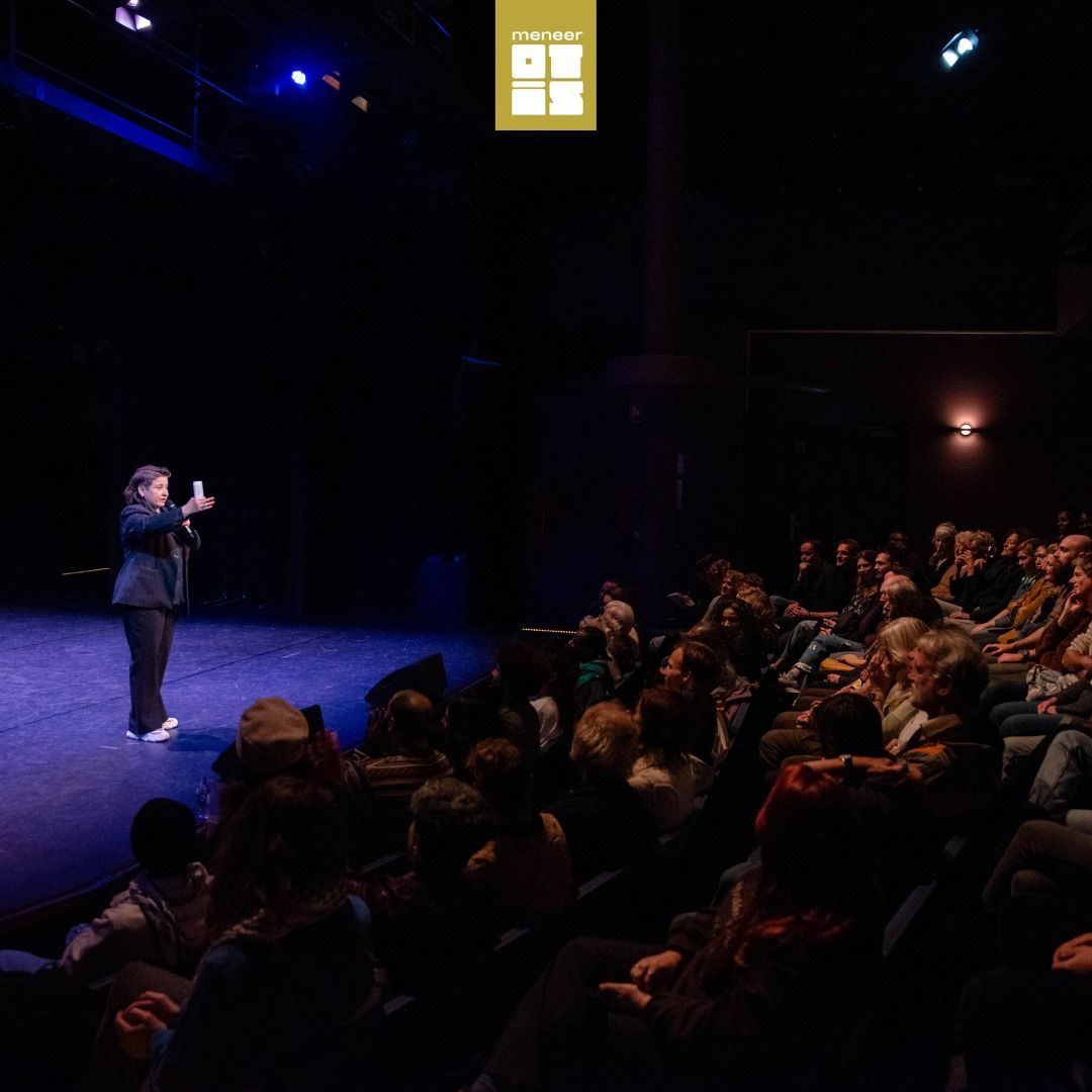 Een spreker staat op een donker podium en spreekt een publiek toe dat in een theater zit onder blauw licht.