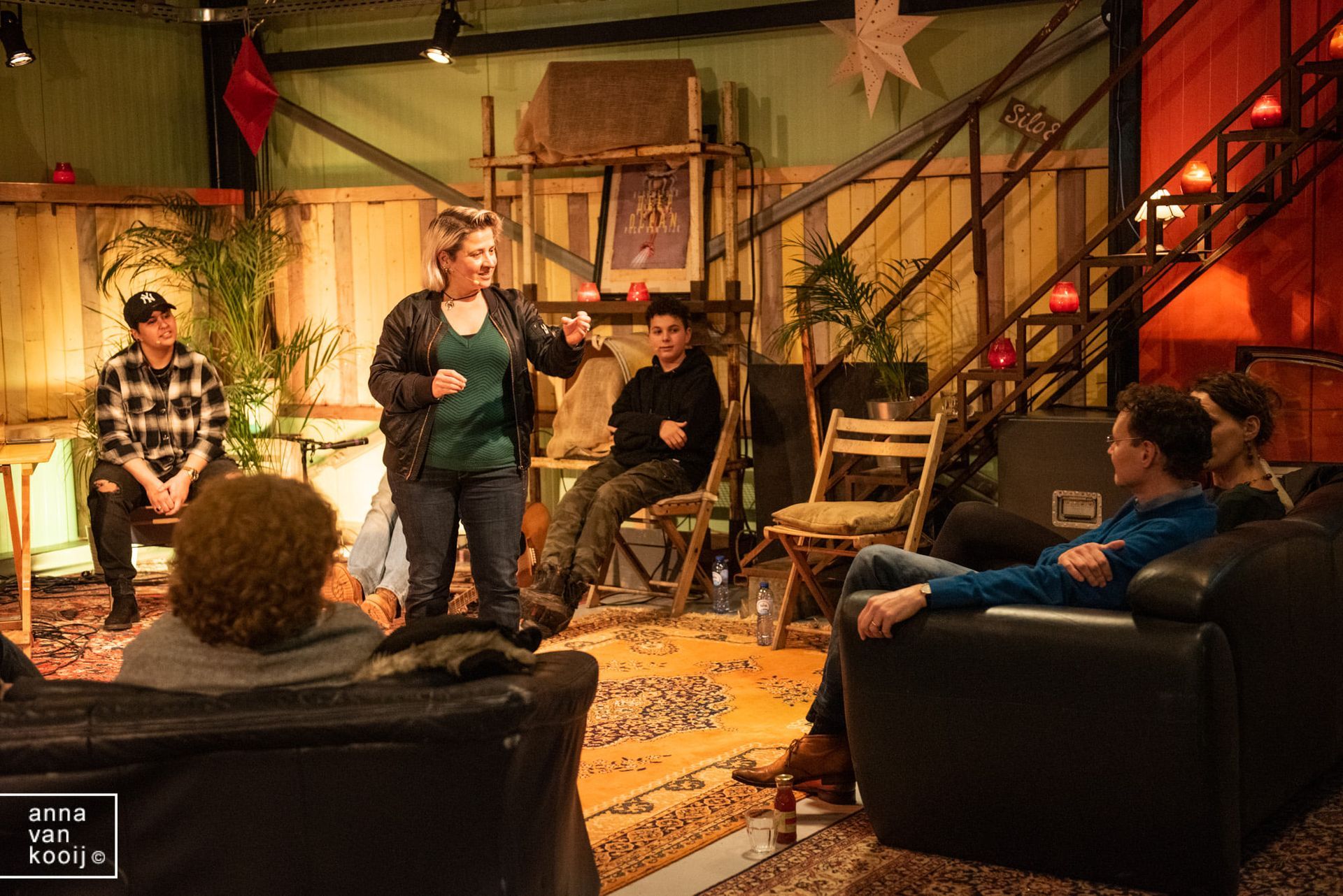 Mensen zitten in een knusse studio-audience en kijken naar een spreker op een sfeervol verlichte set met trappen en stoelen.