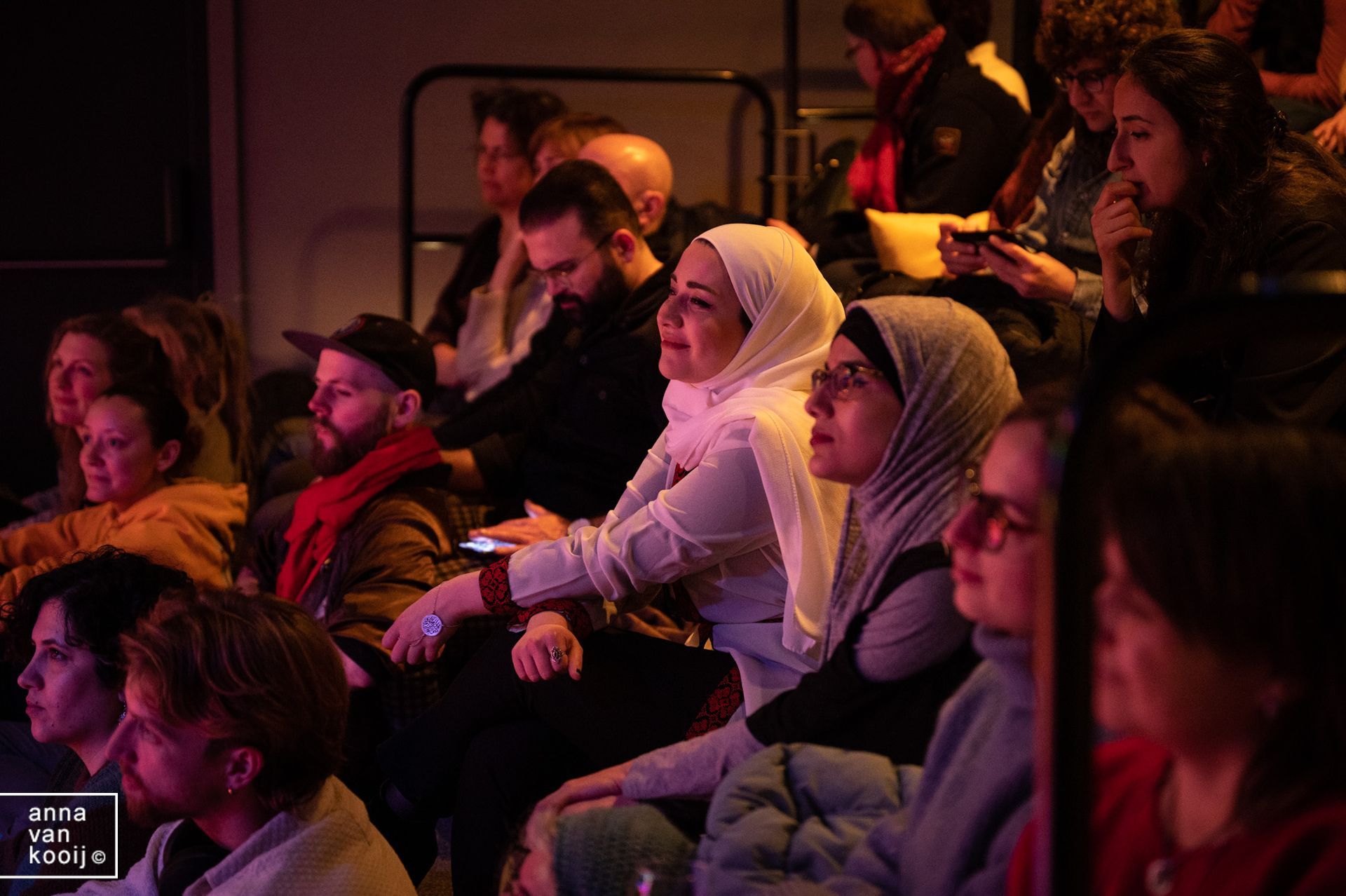 Het publiek zit in een schemerig theater en kijkt naar een voorstelling bij warm podiumlicht.