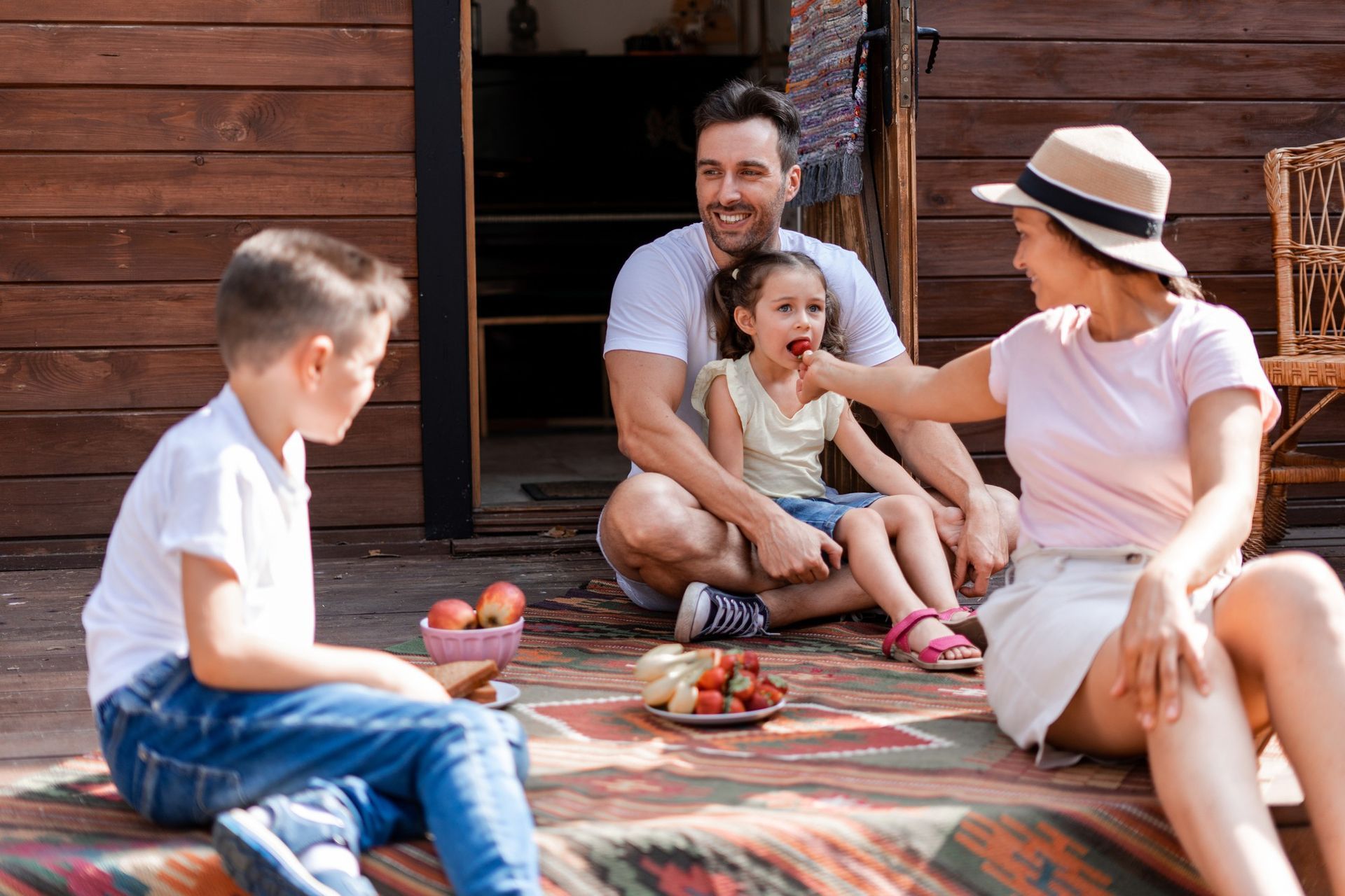 Family enjoying strawberries outdoors: Mother feeds a strawberry to a child, sitting with father and another child.