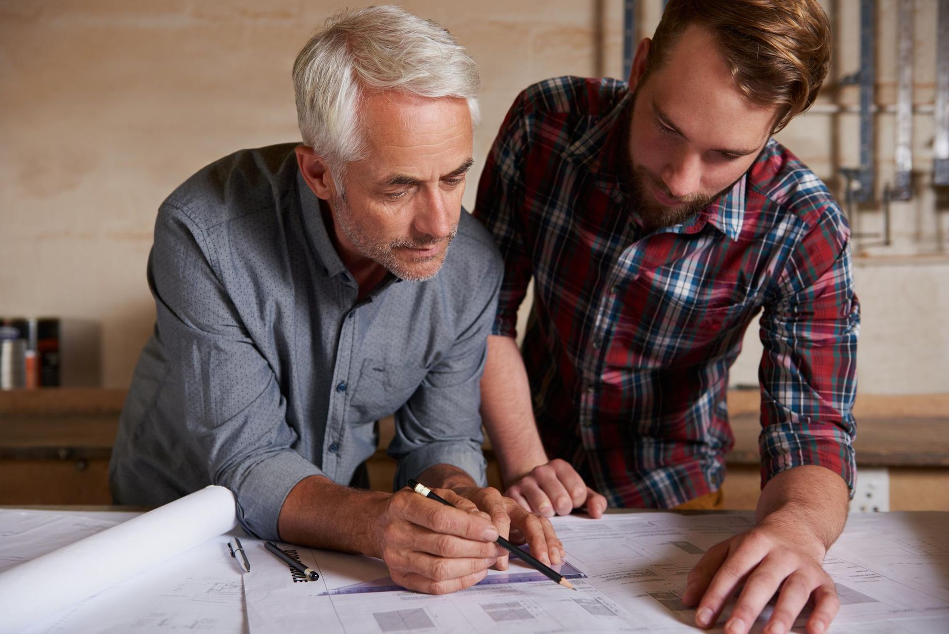 Two men looking at blueprints, one pointing with a pencil. Workshop setting, focused expressions.
