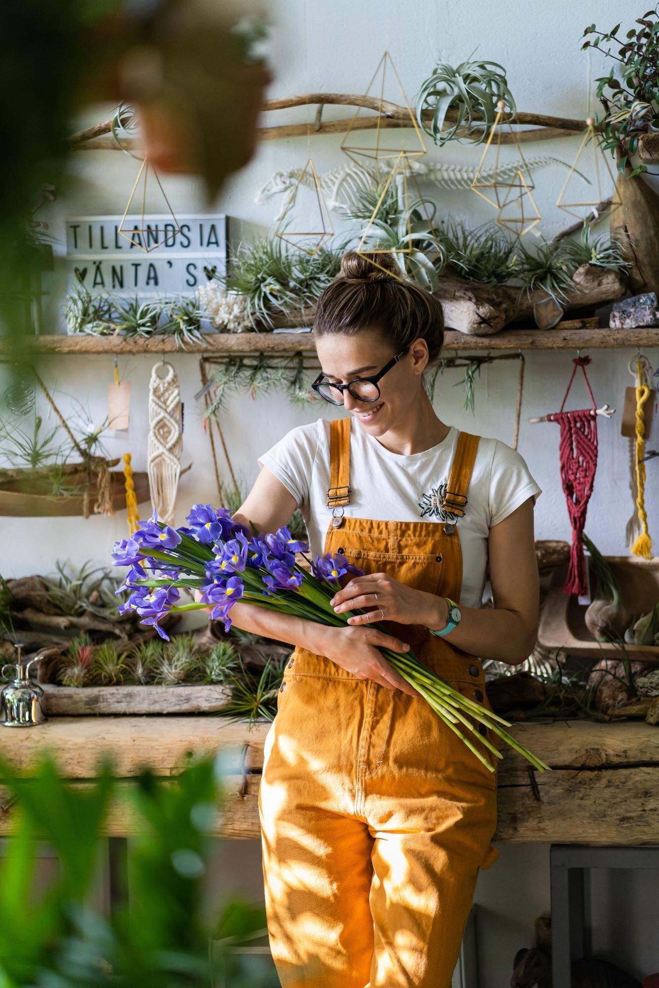 Woman in orange overalls holding purple flowers, smiles in a flower shop.