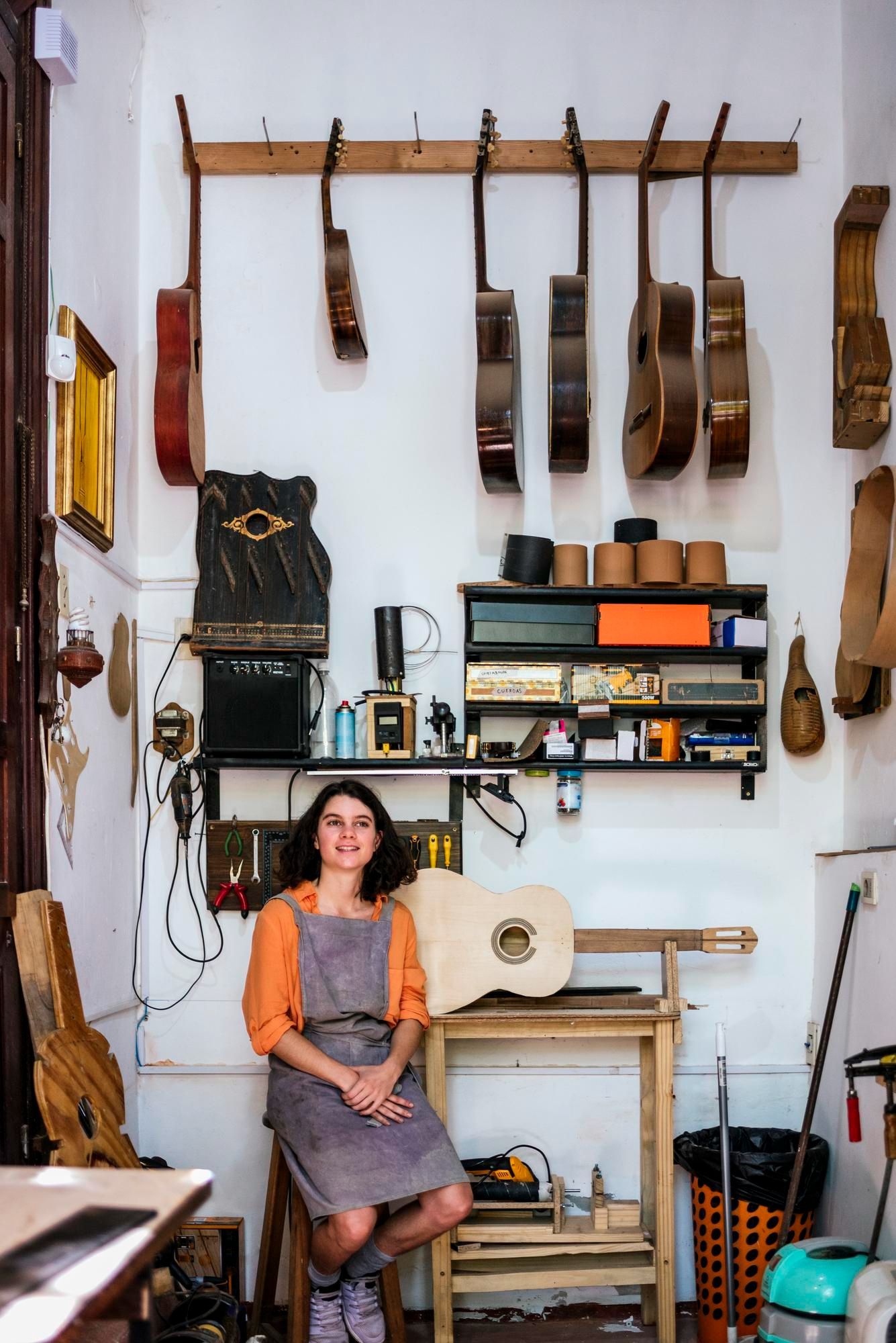 Woman in workshop leans by a guitar. Guitars hang on the wall, tools on shelves.