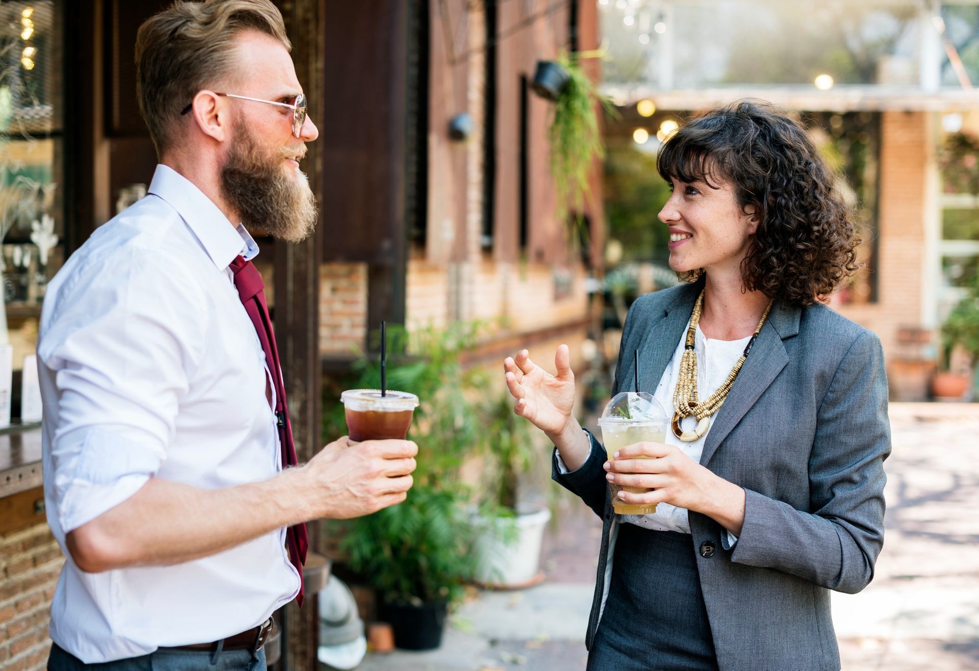 Man with beard and woman in blazer talking and holding iced drinks outdoors.