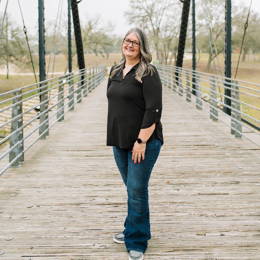 Woman wearing glasses, black shirt, and turquoise pendant smiles in front of a green textured wall.