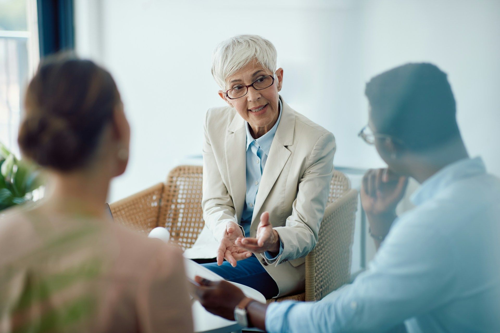 A woman with glasses gestures while talking to two people seated in a room; a discussion is in progress.