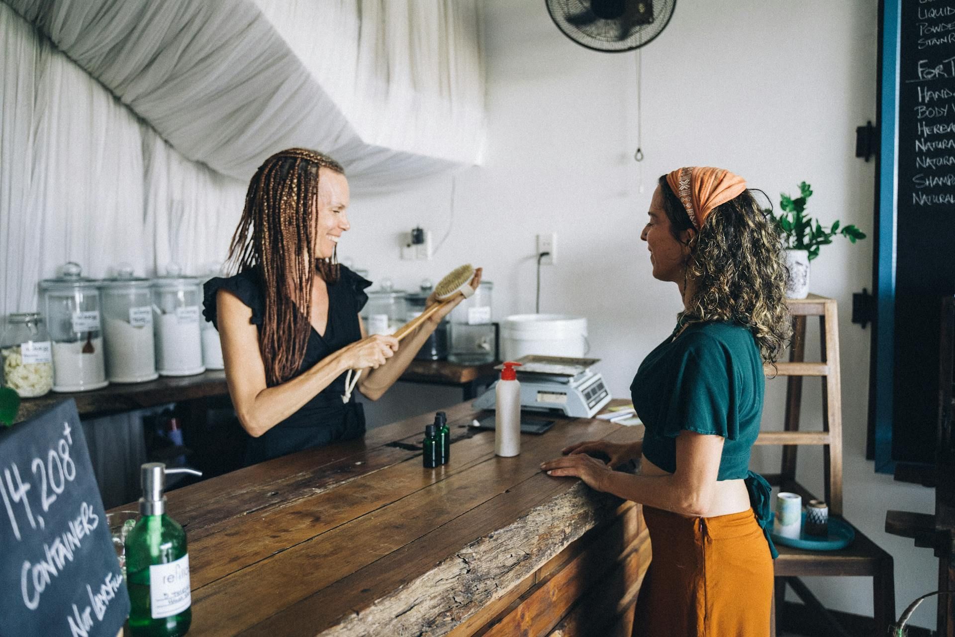 Woman showing a customer a wooden brush at a zero-waste store counter. Jars, scales, and product bottles are visible.