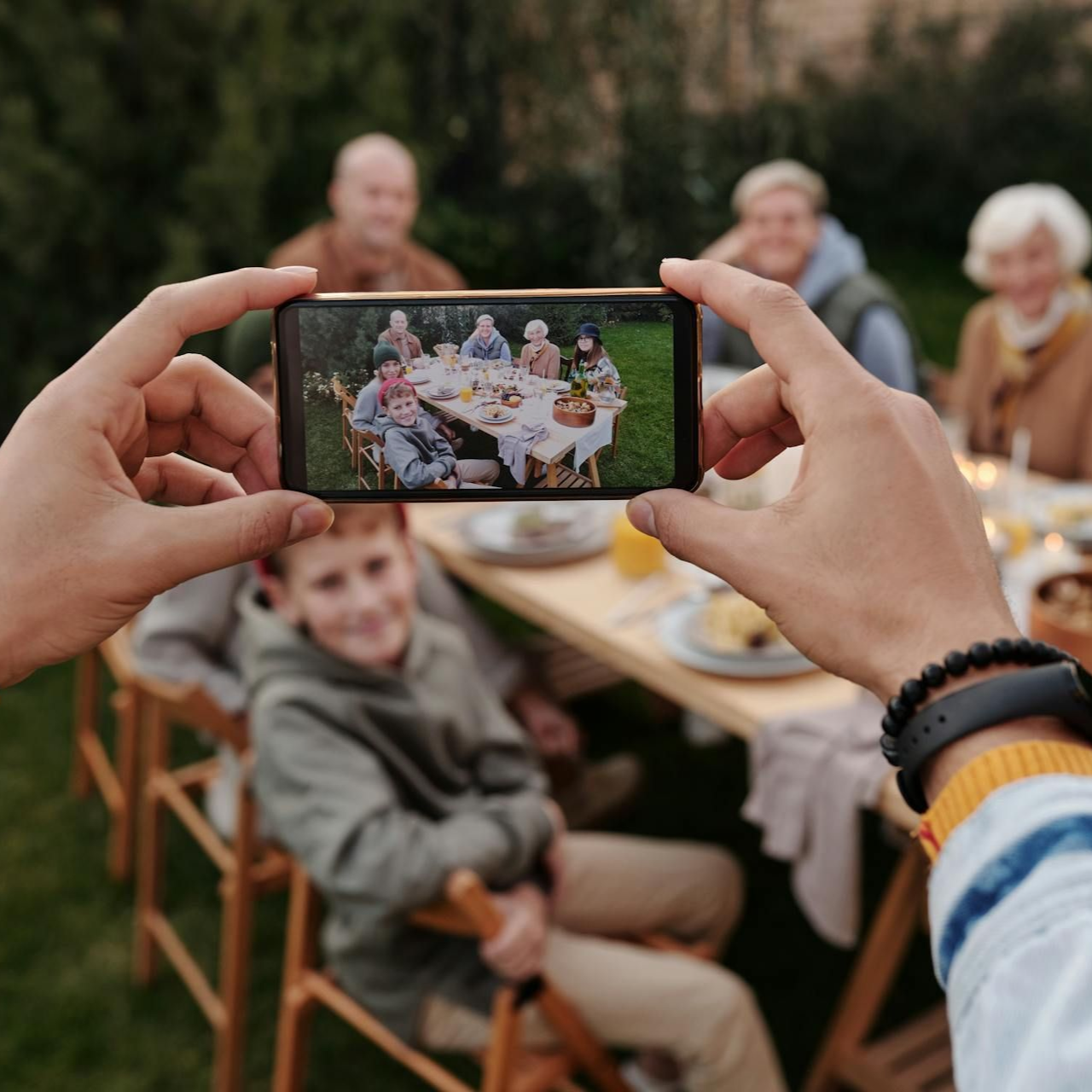 Hands holding phone, taking photo of family seated at outdoor table.