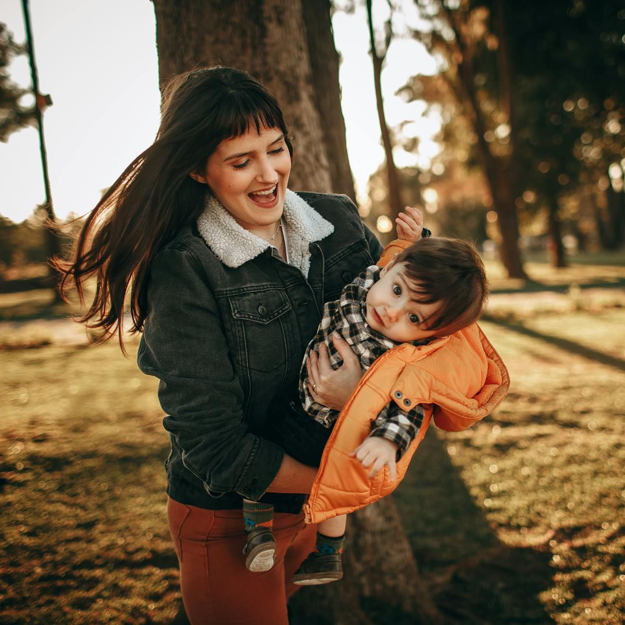 Woman smiling, holding a child wearing an orange jacket in a park, near a tree.