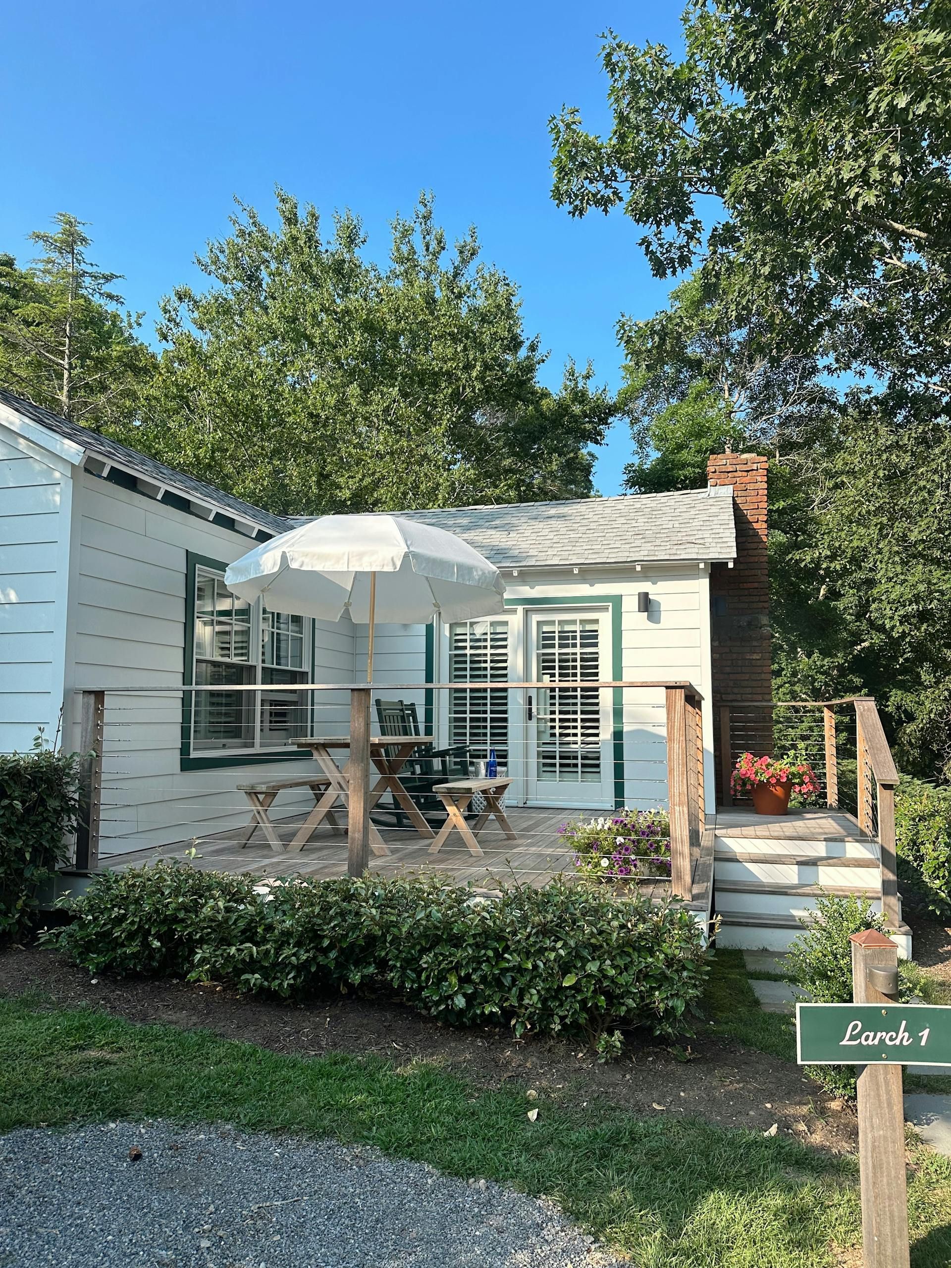 White cabin with small porch and outdoor seating under a white umbrella, surrounded by green trees and foliage.
