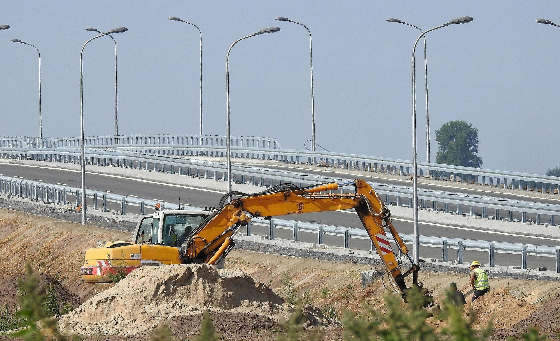 Yellow excavator digging near a bridge with silver guardrails and streetlights, two workers.