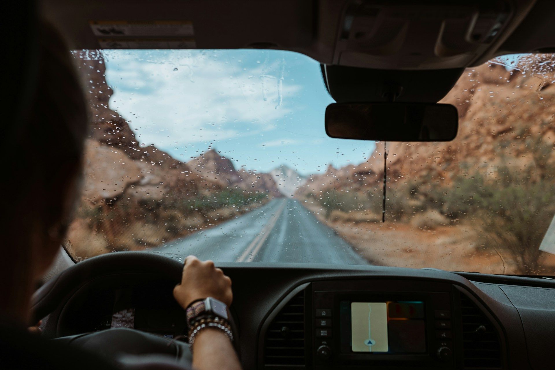 Person driving a vehicle on a desert road, mountains in the distance, windshield covered in rain.