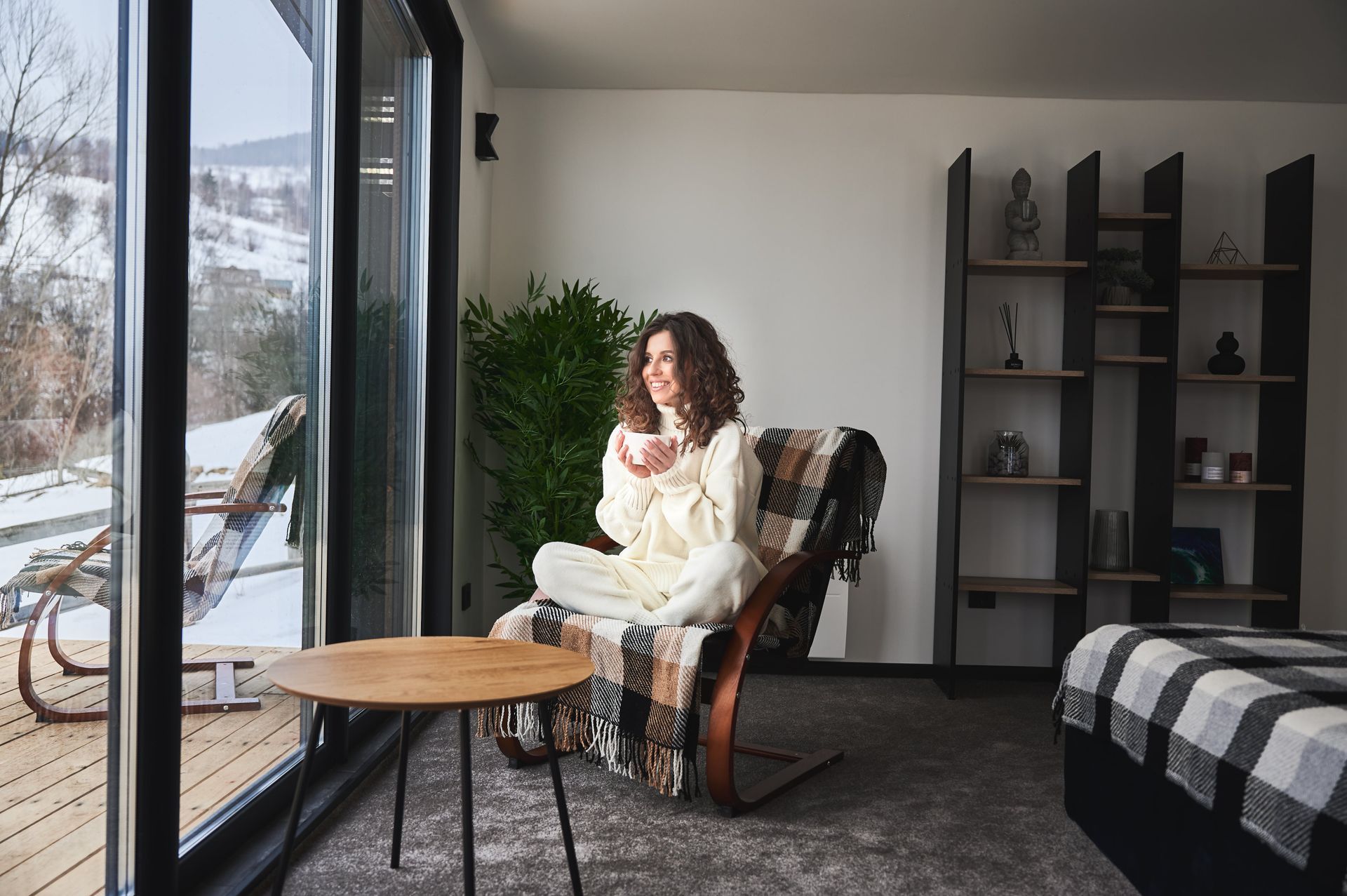 A woman is sitting in a chair in front of a window drinking coffee.
