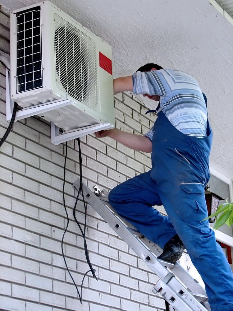A Man Is Standing On A Ladder Fixing An Air Conditioner On A Brick Wall — North West Heating, Cooling and Refrigeration in Moree, NSW