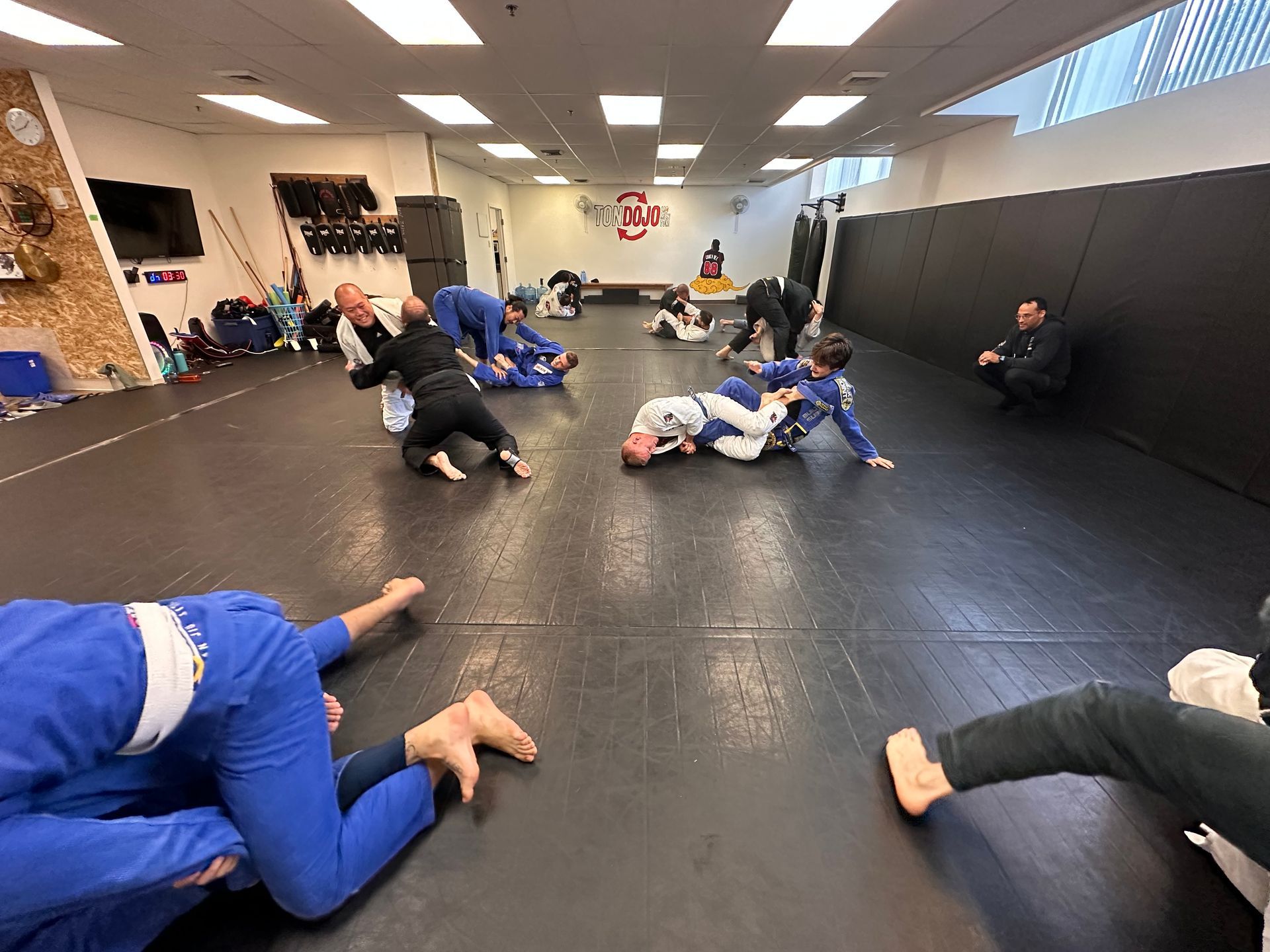 Des pratiquants de jiu-jitsu dans une salle de sport. Plusieurs paires s'affrontent sur des tapis, vêtues de kimonos bleus et noirs.