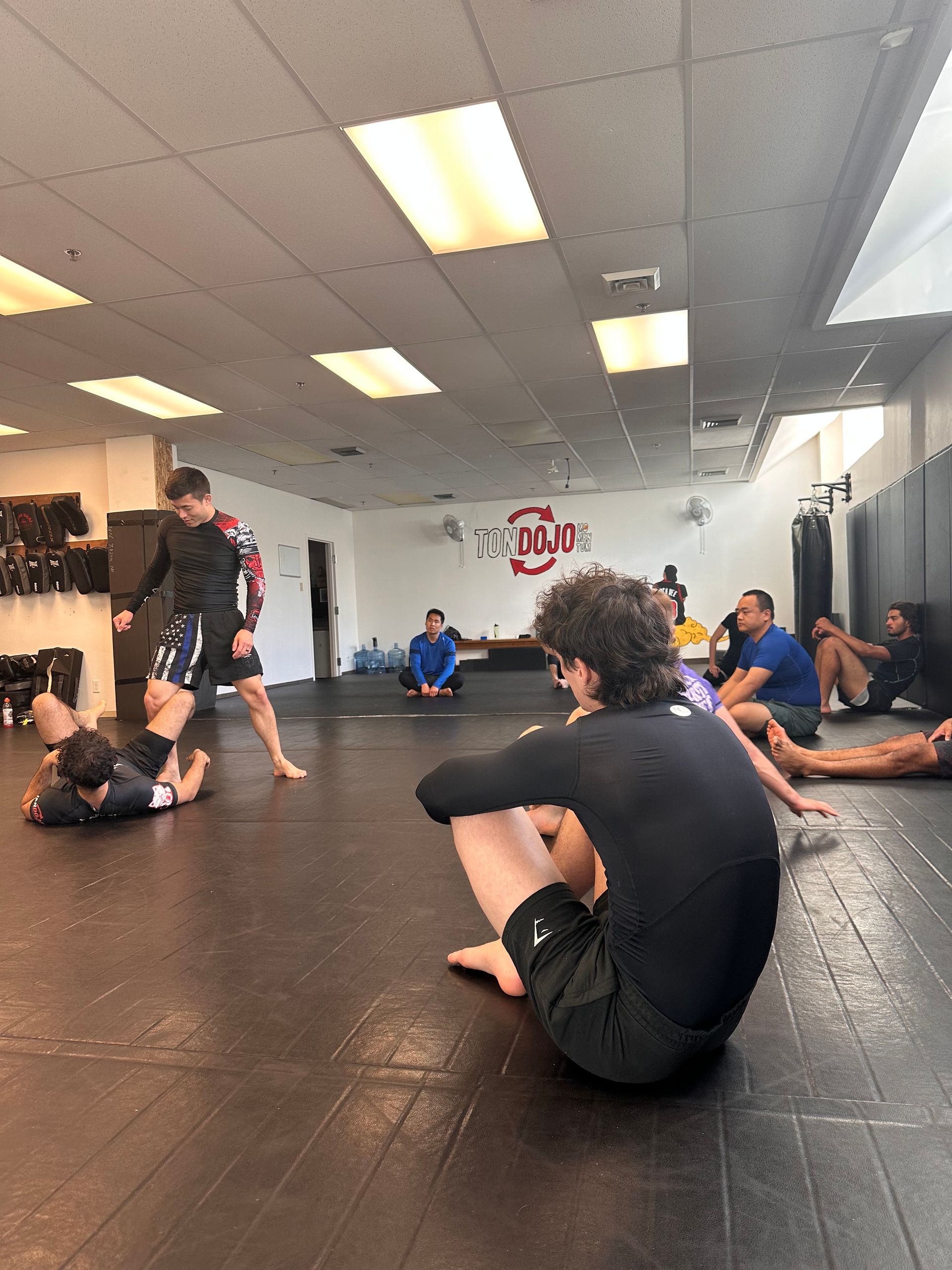 Des personnes participent à un cours de jiu-jitsu. Deux personnes s'affrontent sur un tapis, l'instructeur est debout et les élèves observent depuis une salle de sport.