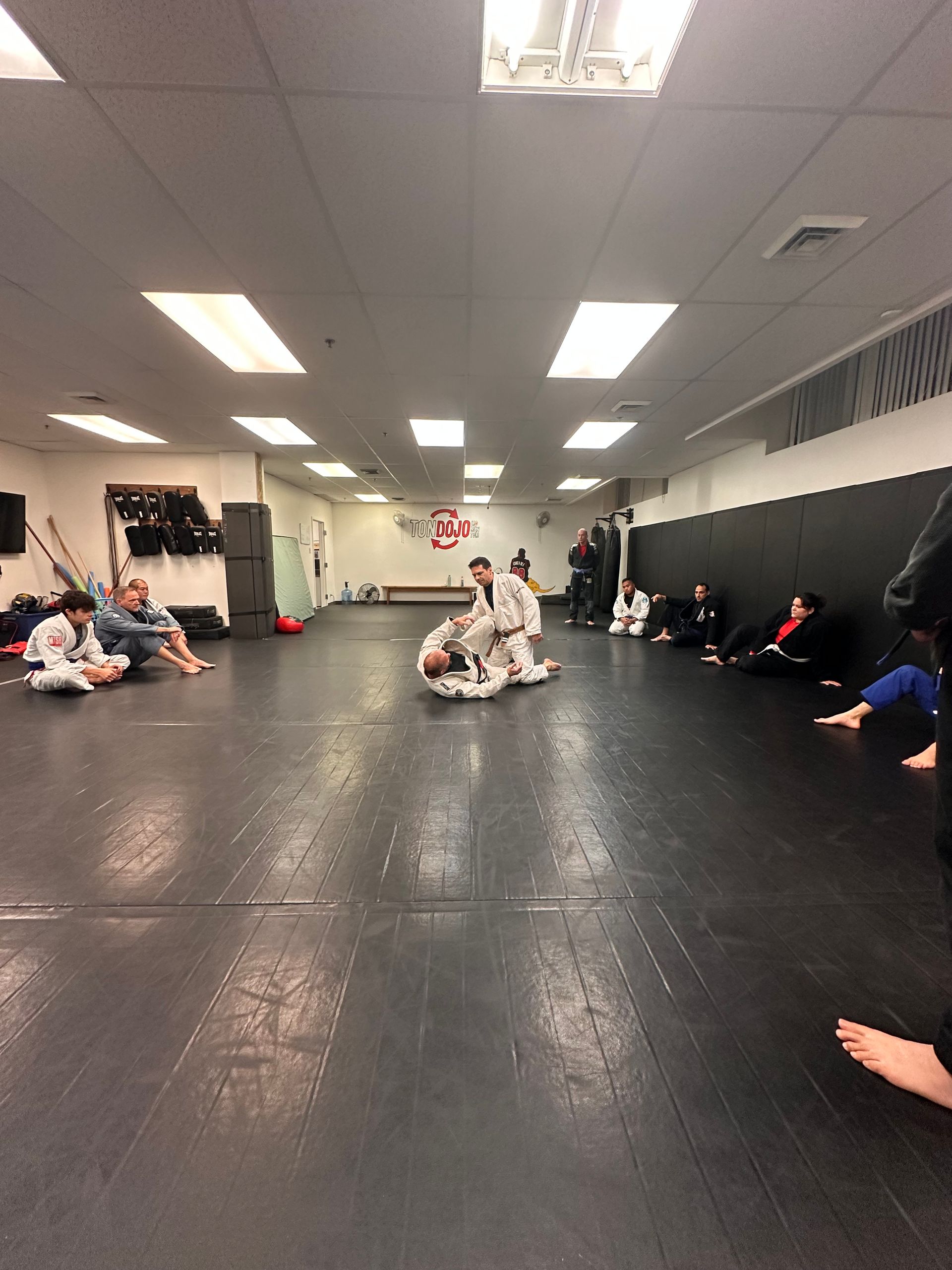 Des personnes participent à un cours d'arts martiaux, s'entraînant sur un tapis noir. Certains sont assis, d'autres luttent. La salle est éclairée par des néons.