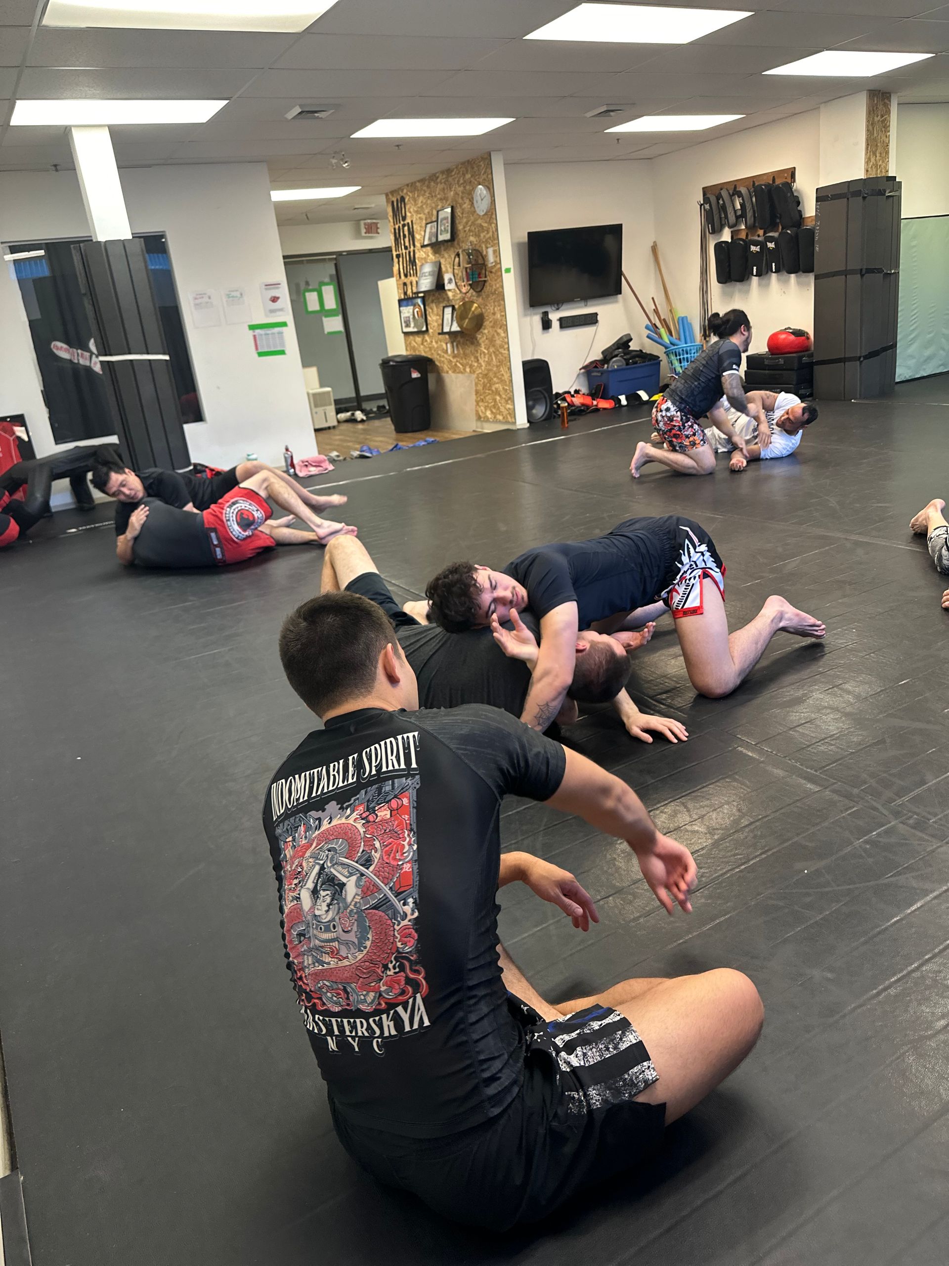 Des pratiquants de Jiu-Jitsu sur un tapis noir dans une salle de sport. Certains luttent, d'autres sont assis.