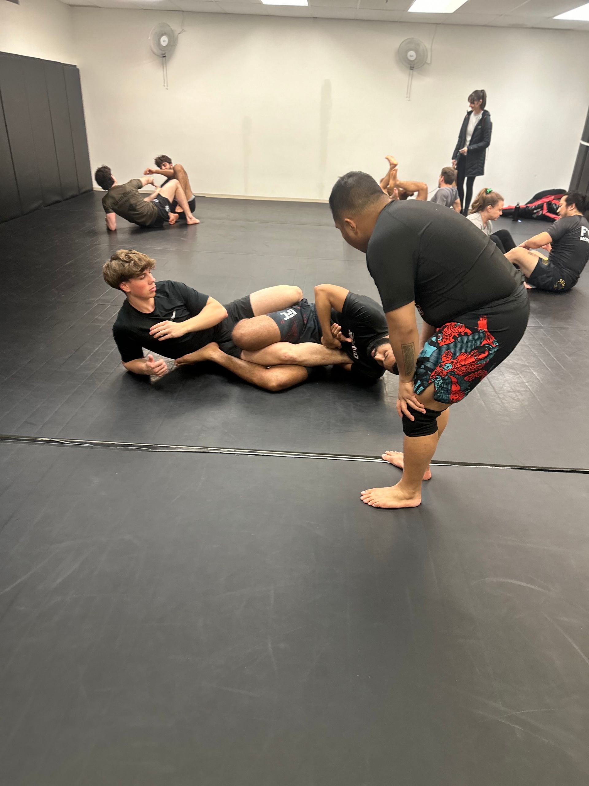 Cours d'arts martiaux : un homme entraîne deux élèves au grappling sur un tapis. D'autres s'entraînent dans une salle de sport.