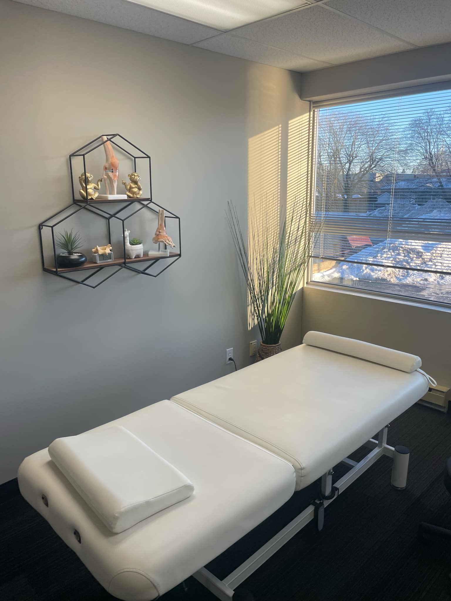 Table de massage dans une pièce bien éclairée, à côté d'une fenêtre, avec une étagère murale décorative au-dessus.