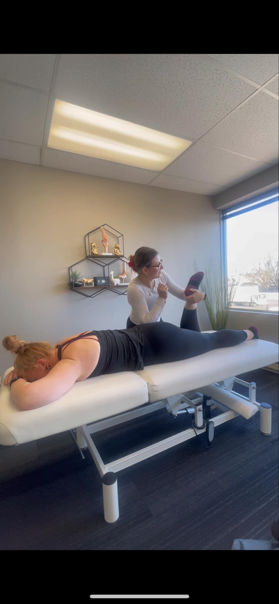 Une femme est allongée sur une table de massage tandis qu'une autre femme soulève sa jambe. Dans un bureau avec fenêtre.