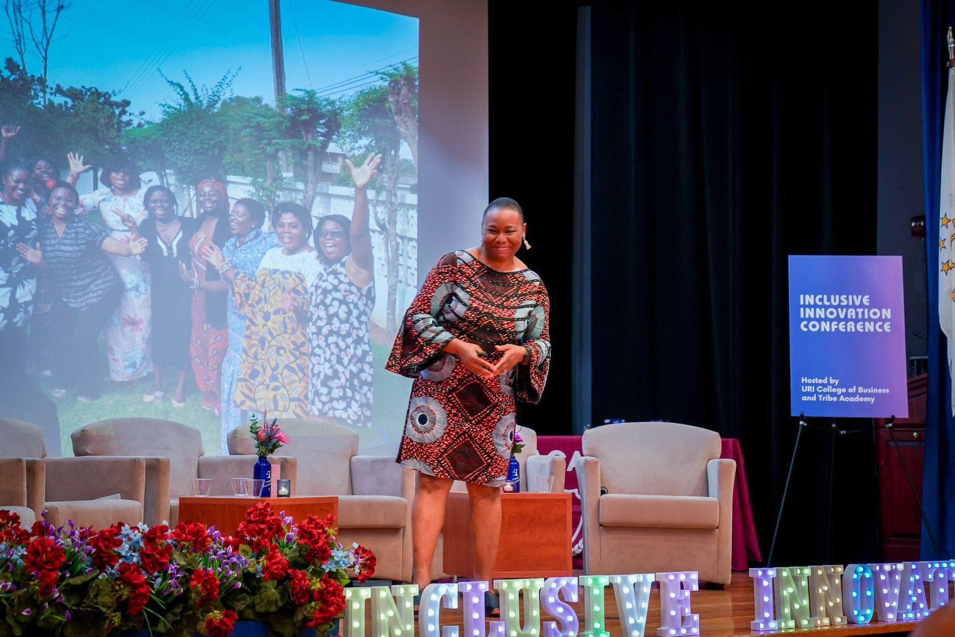 Woman in African print dress speaks on stage; presentation backdrop.