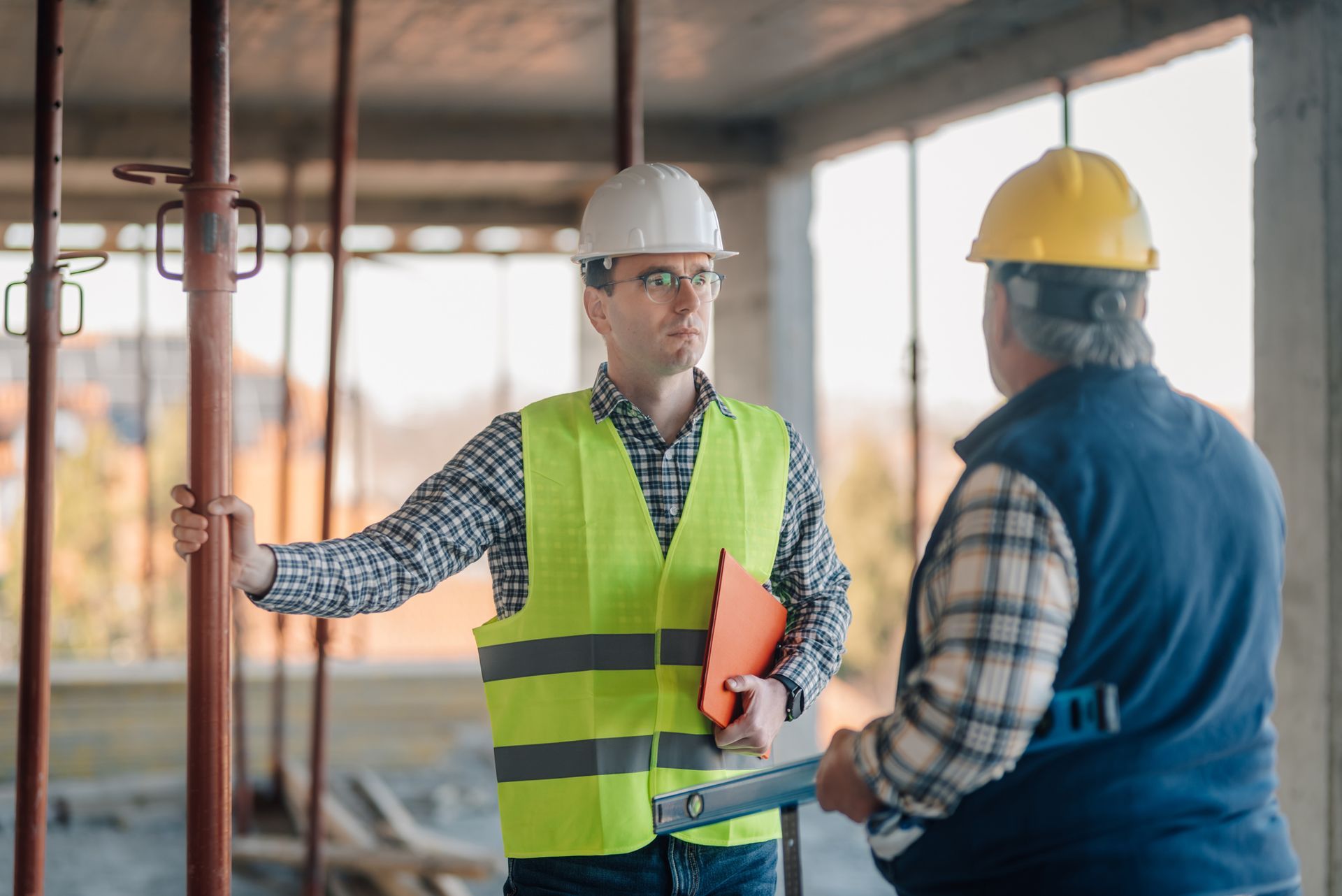 Two engineers wearing safety vests and hardhats are discussing building progress at a construction. Two engineers wearing safety vests and hardhats are discussing building progress at a construction.
