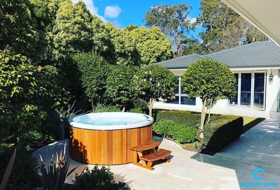 A sunlit backyard features a round wooden hot tub with clear water, accessed by small wooden steps, set on a paved patio. The patio is surrounded by neatly trimmed hedges and sculpted trees. In the background, a single-story house with large glass doors and a light-colored roof overlooks the garden, with dense green foliage and tall trees beyond. A small “Sapphire Spas” logo appears in the lower right corner.
Provide your feedback on BizChat