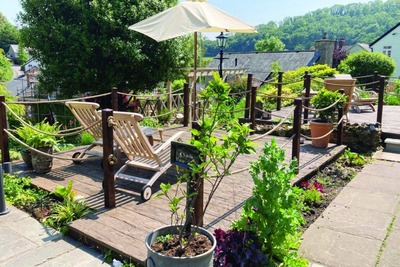Outdoor seating area with wooden lounge chairs, umbrella, and rope fencing overlooking a village.