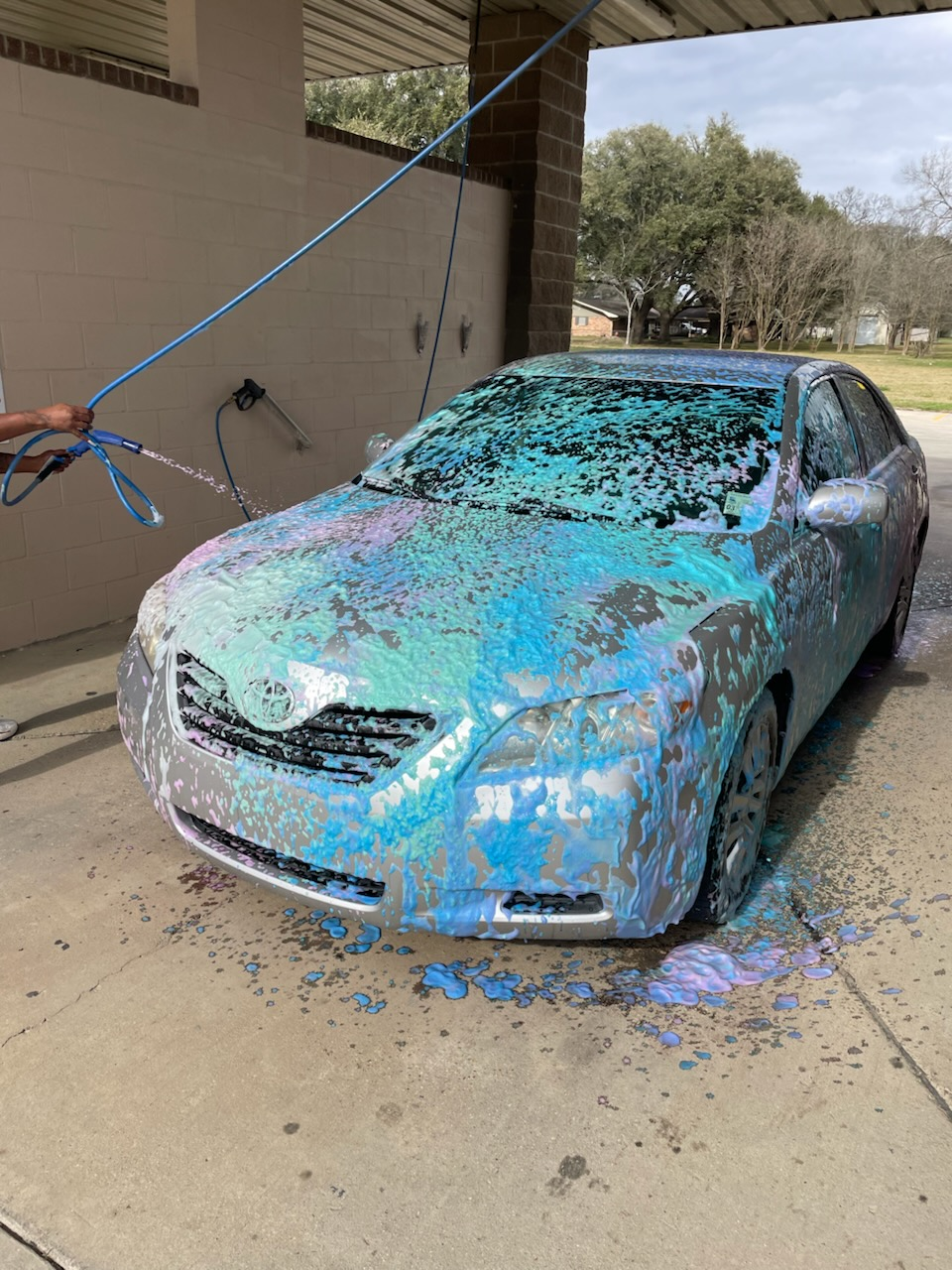 A silver sedan covered in thick blue and purple suds being washed at an outdoor car wash station.