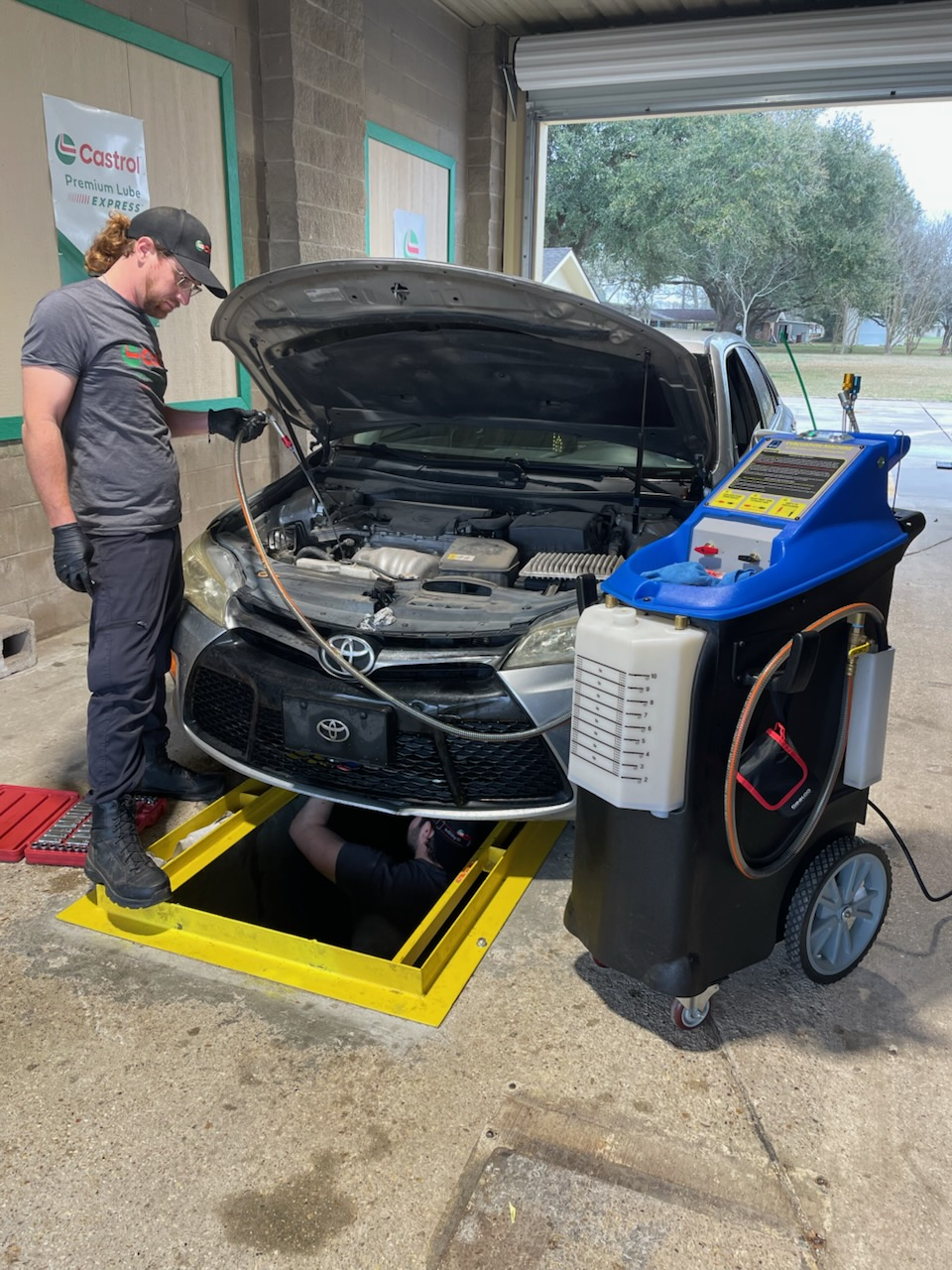 A technician services a Toyota car over an open service pit, with a blue fluid exchange machine in the foreground.