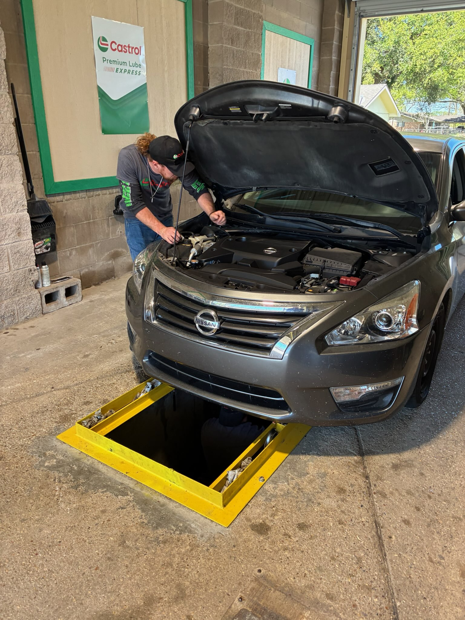 A mechanic works on a grey sedan positioned over an open yellow service pit in a garage.