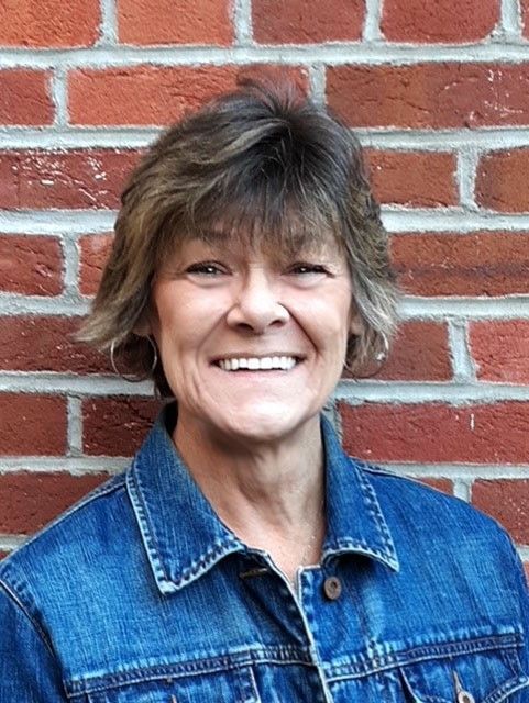 Smiling person in a blue denim jacket standing in front of a red brick wall