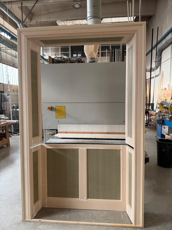 Finestra con telaio in legno e vetro in un'officina, con una sezione inferiore a pannelli e macchinari alle sue spalle.
