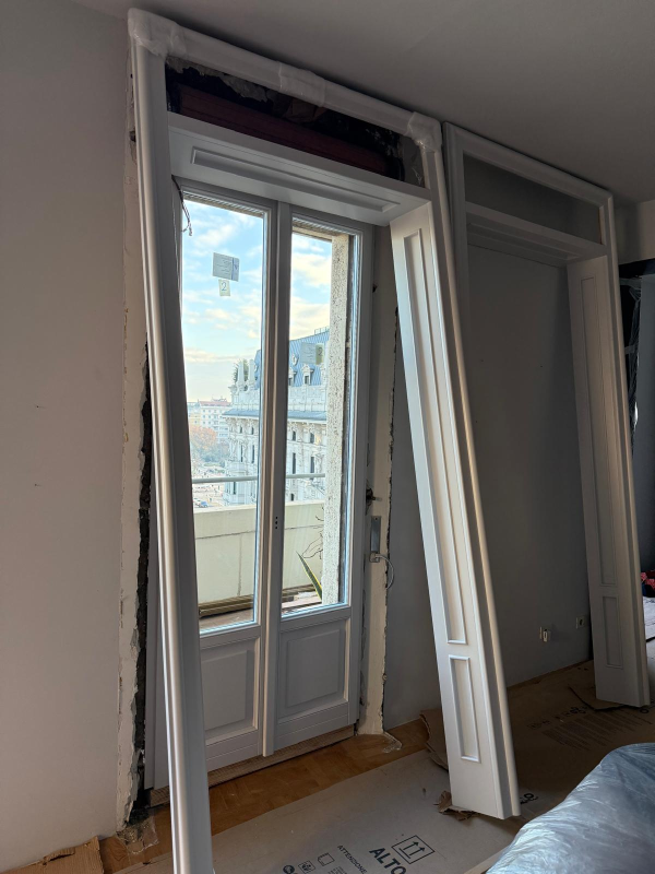Porta finestra aperta con cornice bianca e vista sulla città; materiali per la ristrutturazione degli interni sono appoggiati nelle vicinanze.