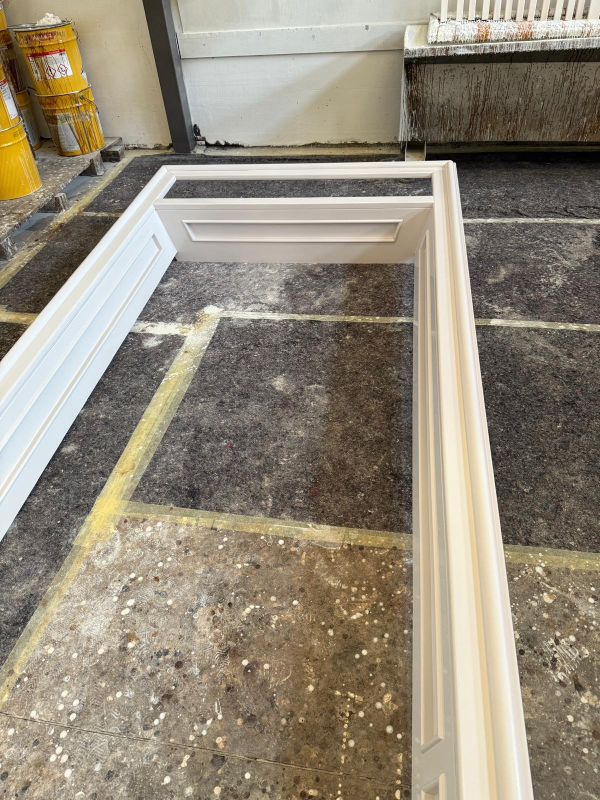 Telaio bianco di una finestra appoggiato sul pavimento di un'officina, accanto a un muro e a dei bidoni della spazzatura.