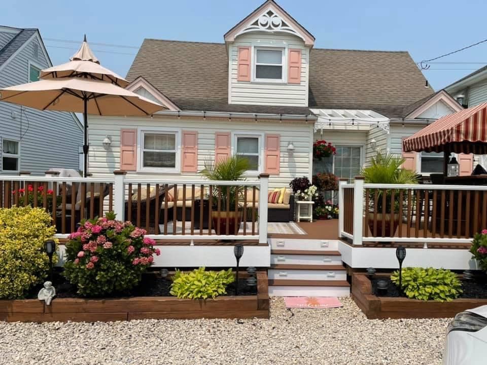 Front view of a pastel cottage with white porch, flower boxes, and patio umbrellas in a landscaped yard