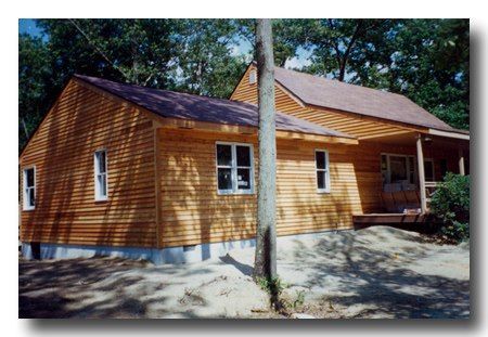 Wooden cabin with a front porch in a wooded area, seen from the front-left side.