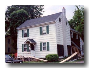 White two-story house with green shutters and a small front porch, leaning heavily to one side