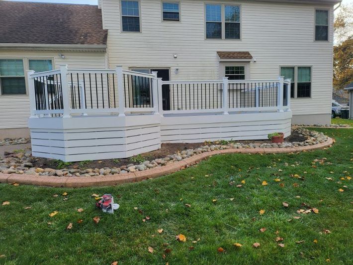 White raised deck with railings attached to a beige house, bordered by a stone bed and green lawn.