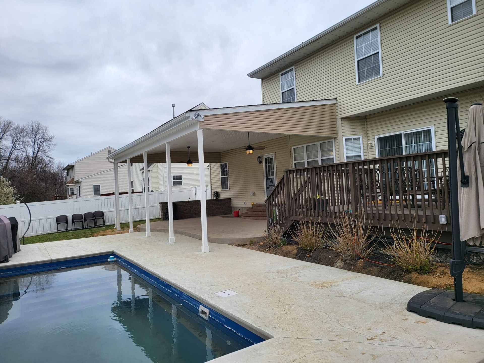 Backyard pool beside a house with a white-covered patio and wooden deck under a cloudy sky