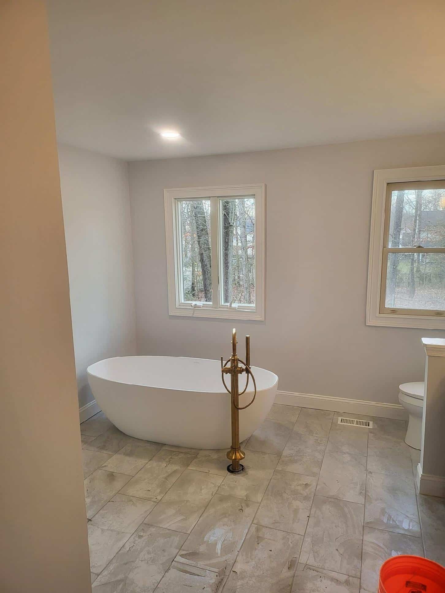 Bright bathroom with freestanding tub, marble floor, and two windows overlooking trees.