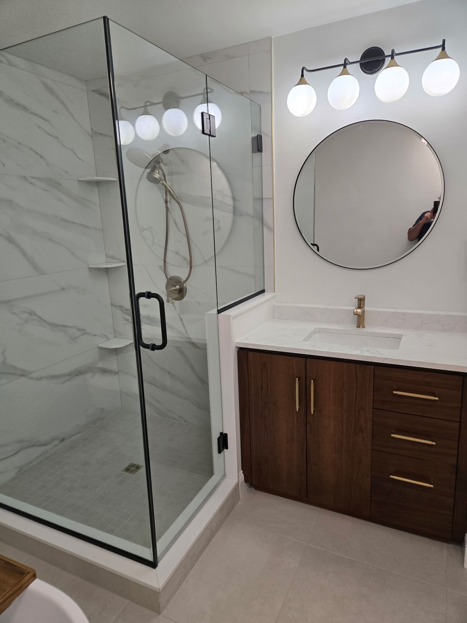 Modern bathroom with glass shower, round mirror, vanity, and lit three-bulb vanity lights