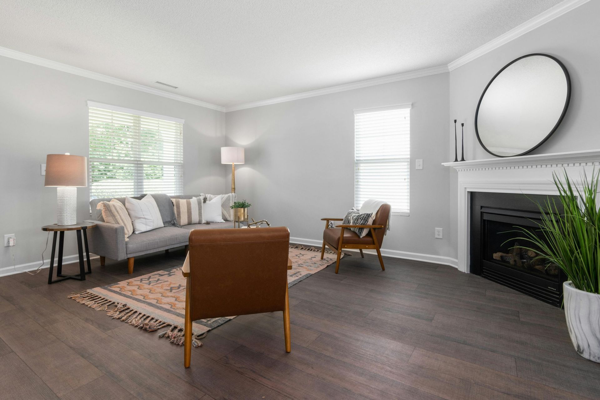 Bright living room with gray sofa, wood floors, round mirror above fireplace, and tall plants