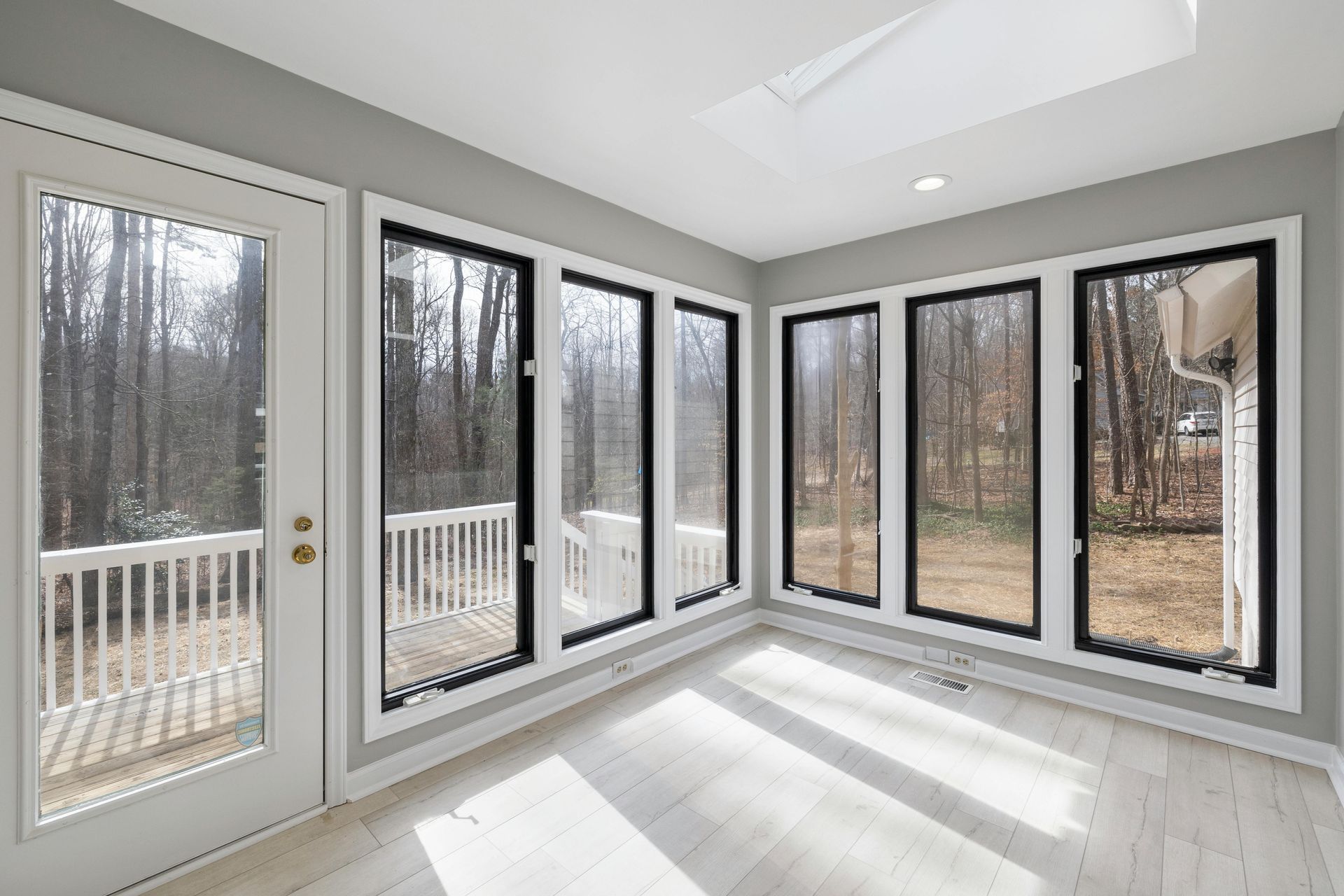 Bright sunroom with tall black-trimmed windows and glass door overlooking a wooded deck