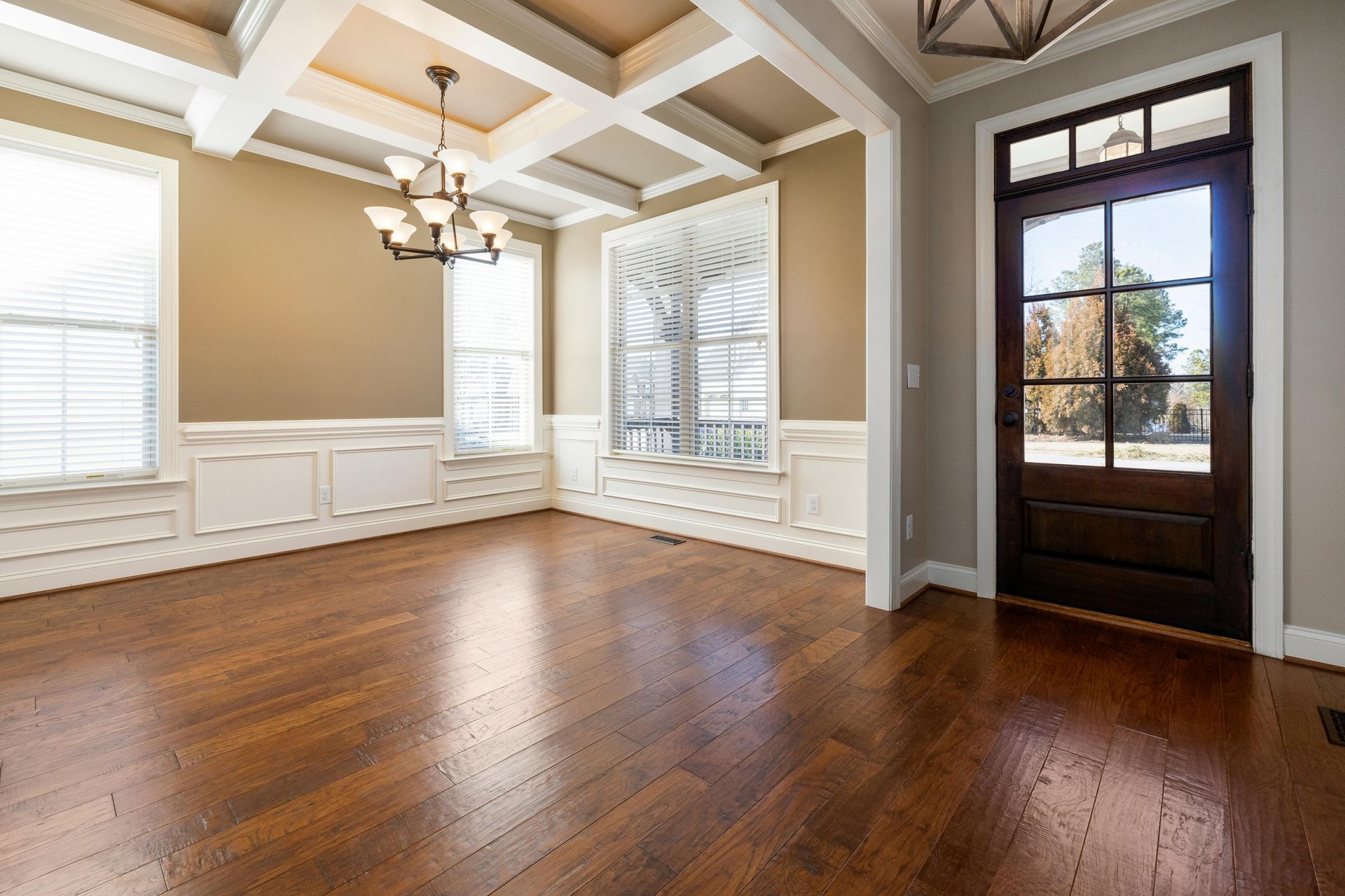 Empty dining room with hardwood floors, beige walls, white trim, chandelier, and dark front door