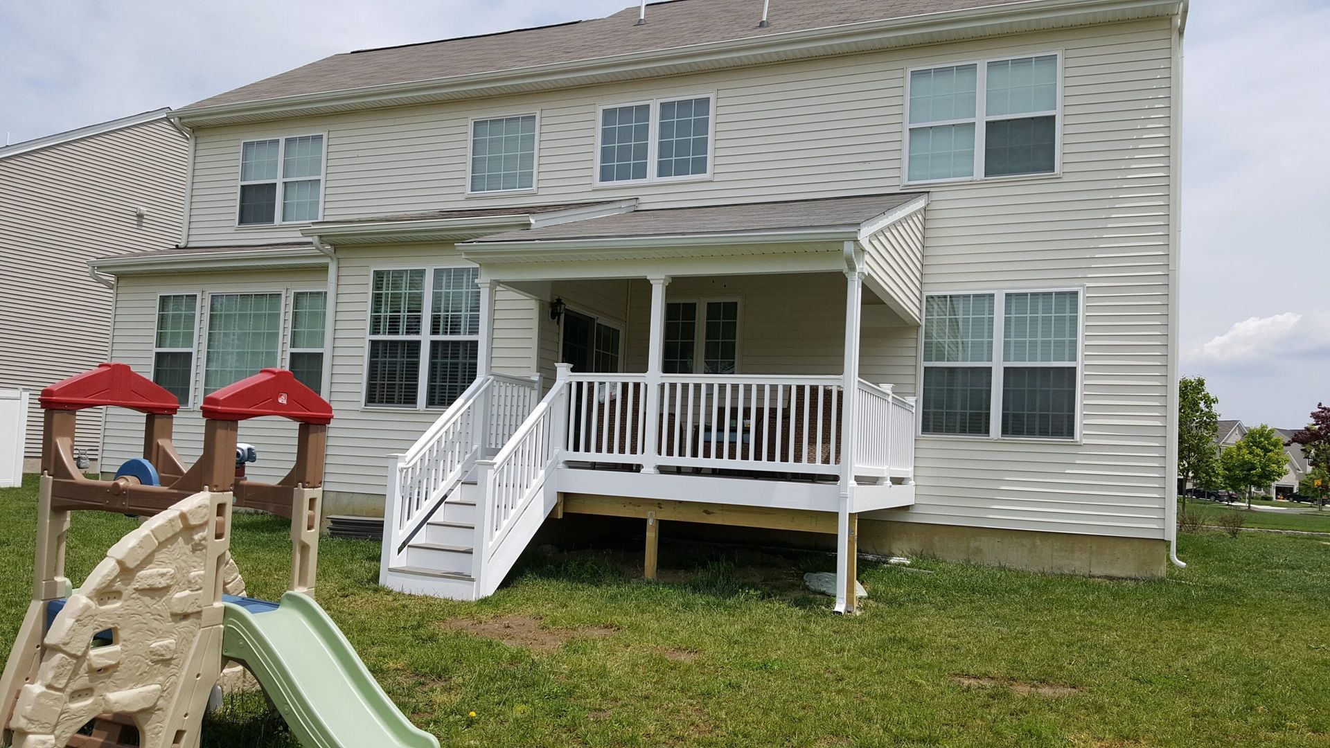 Backyard view of a two-story house with a white deck and stairs, red patio umbrellas, and a children’s slide